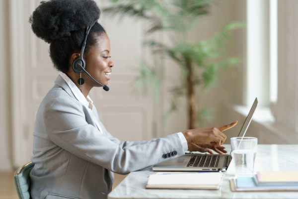 Smiling African employee woman with headphones pointing finger on laptop screen, working at laptop at home office, talking in video chat with clients, consulting customer, distance webinar online for six sigma.